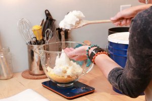 Jess adding coconut oil to a glass measuring bowl that is sitting on a digital scale and already contains beeswax.